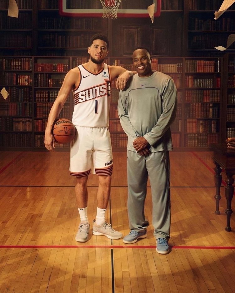 Two adult men pose on a basketball court in front of bookshelves; one man wears a Suns jersey and holds a basketball.