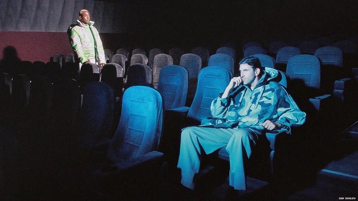 Two adult men in an empty movie theatre; one wearing a camo hoodie seated, and another standing in the background wearing a puffy jacket.