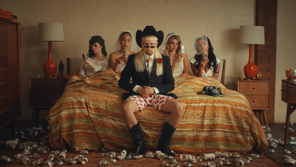Man in a black suit and cowboy hat sits on a bed surrounded by four women in wedding dresses, with cans scattered on the floor.