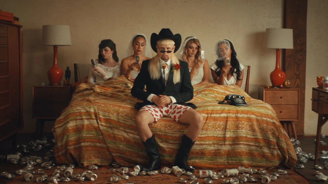 Man in a black suit and cowboy hat sits on a bed surrounded by four women in wedding dresses, with cans scattered on the floor.
