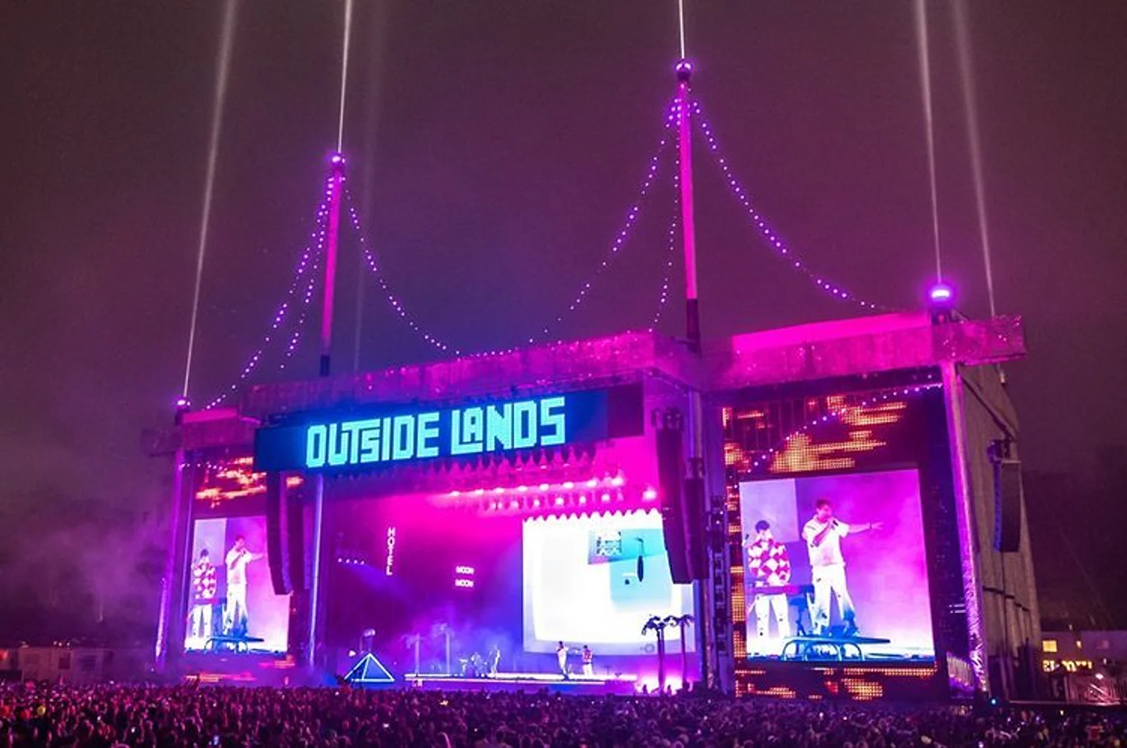 Large crowd gathers at night to watch musicians perform on a huge stage with bright screens at the Outside Lands music festival.