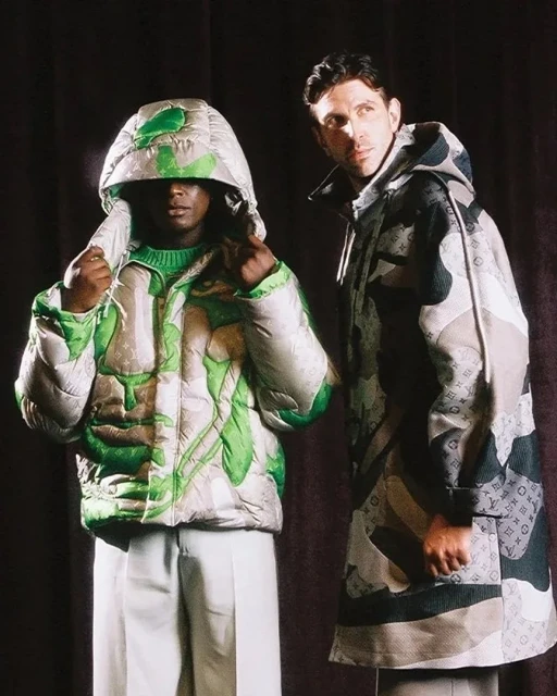 Two fashion models pose in a photo studio, showcasing patterned hooded puffer jackets and light pants against a dark fabric backdrop.