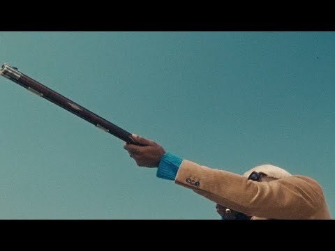 Man in a beige jacket aiming a vintage rifle against a clear blue sky, blending fashion with antique weaponry in a striking outdoor scene.