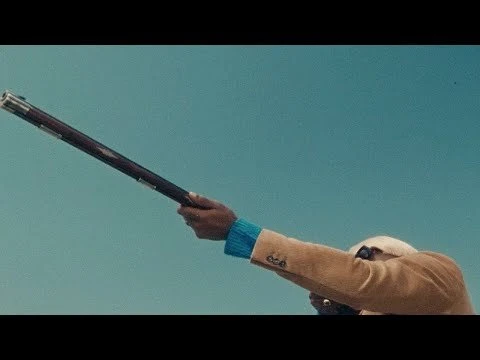 Man in a beige jacket aiming a vintage rifle against a clear blue sky, blending fashion with antique weaponry in a striking outdoor scene.