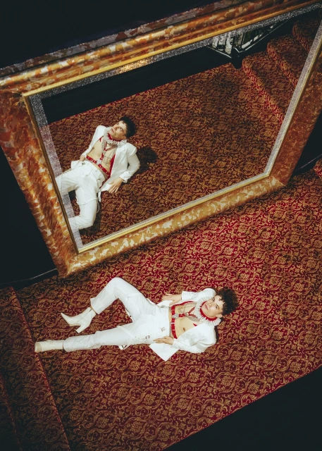 Overhead studio shot of a young adult man in a white suit and white boots lying on a red patterned carpet while framed by a mirror.