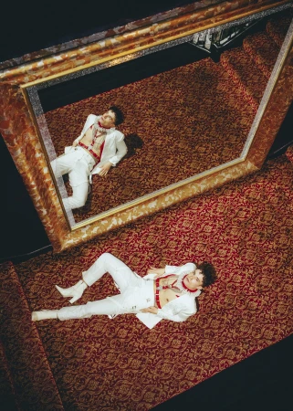 Overhead studio shot of a young adult man in a white suit and white boots lying on a red patterned carpet while framed by a mirror.
