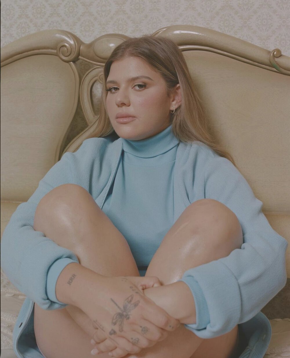 Portrait of a woman with light brown hair in a light blue sweater sitting on a vintage bed with a patterned headboard.