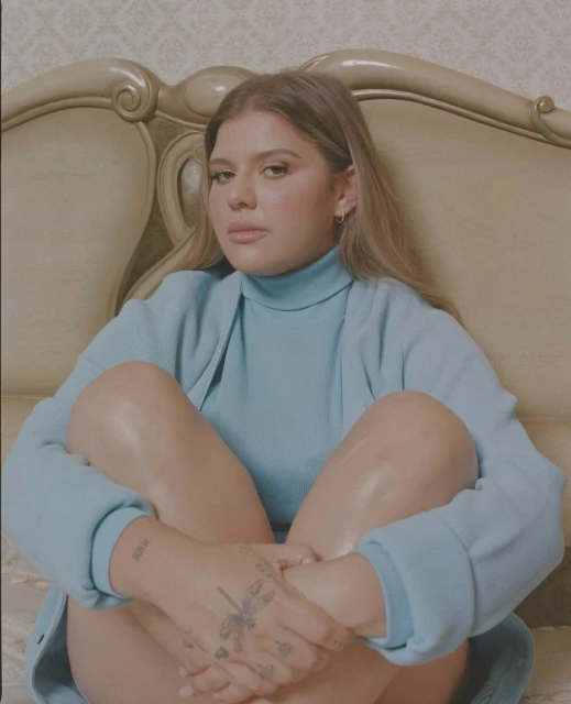 Portrait of a woman with light brown hair in a light blue sweater sitting on a vintage bed with a patterned headboard.