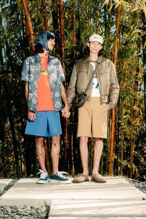 Two men in casual summer fashion hold hands outdoors surrounded by bamboo; one in a headscarf, the other in a baseball cap.