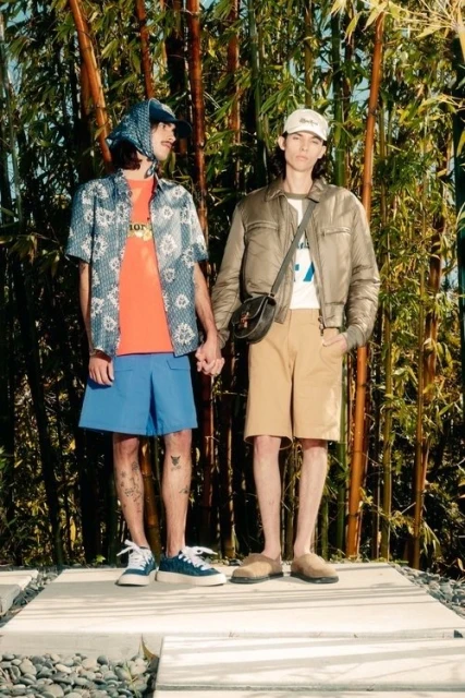 Two men in casual summer fashion hold hands outdoors surrounded by bamboo; one in a headscarf, the other in a baseball cap.
