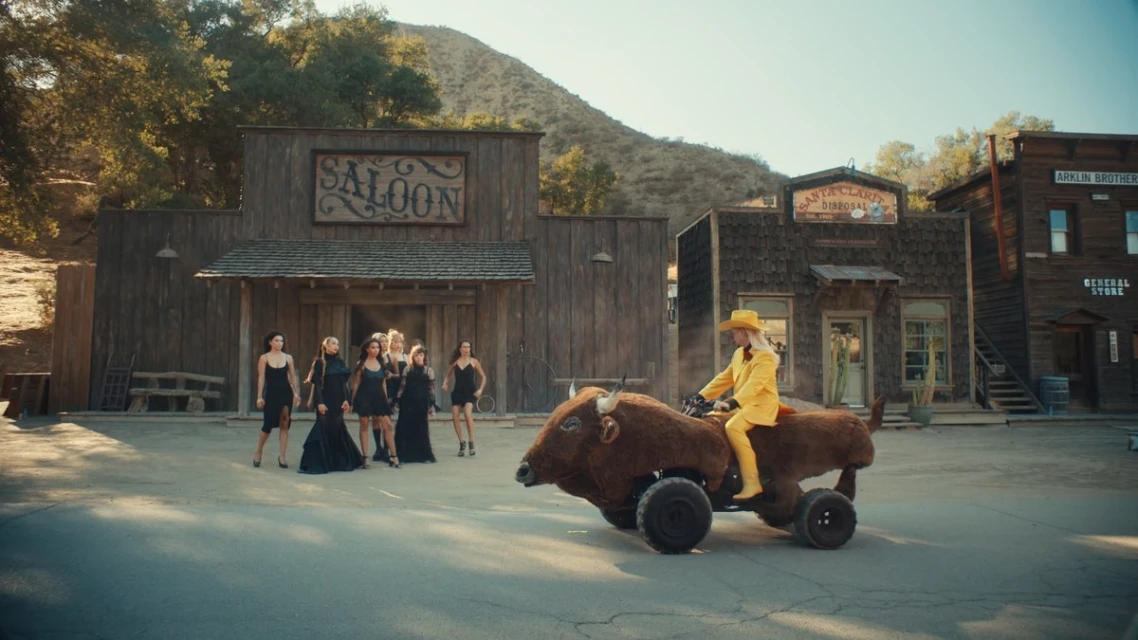 A woman rides a faux buffalo car in a Western town with a group of women in black dresses standing near a wooden 'Saloon' building.