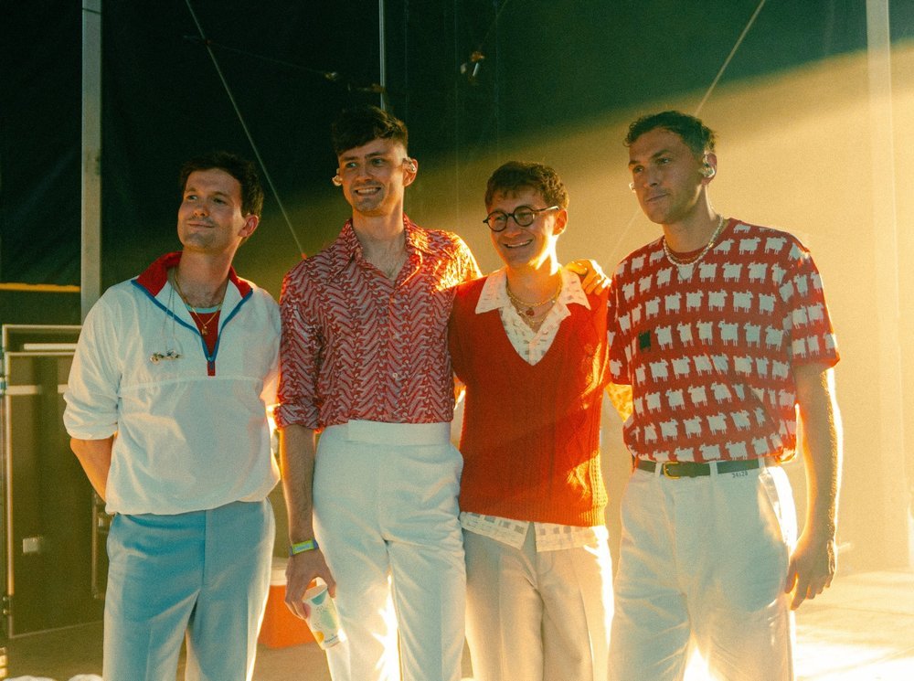 A posed group portrait of four young adult men in retro-inspired clothing against a brightly lit background in a stage-like setting.