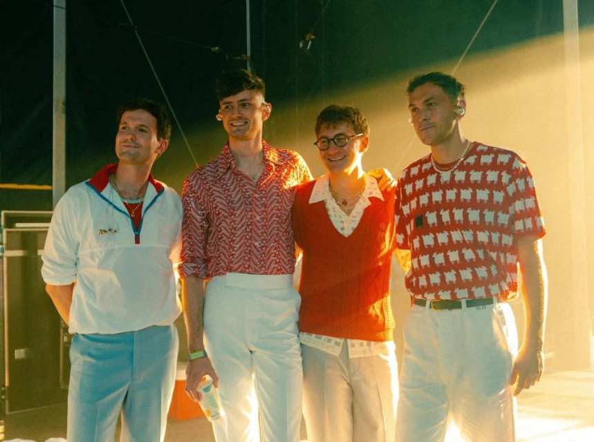 A posed group portrait of four young adult men in retro-inspired clothing against a brightly lit background in a stage-like setting.