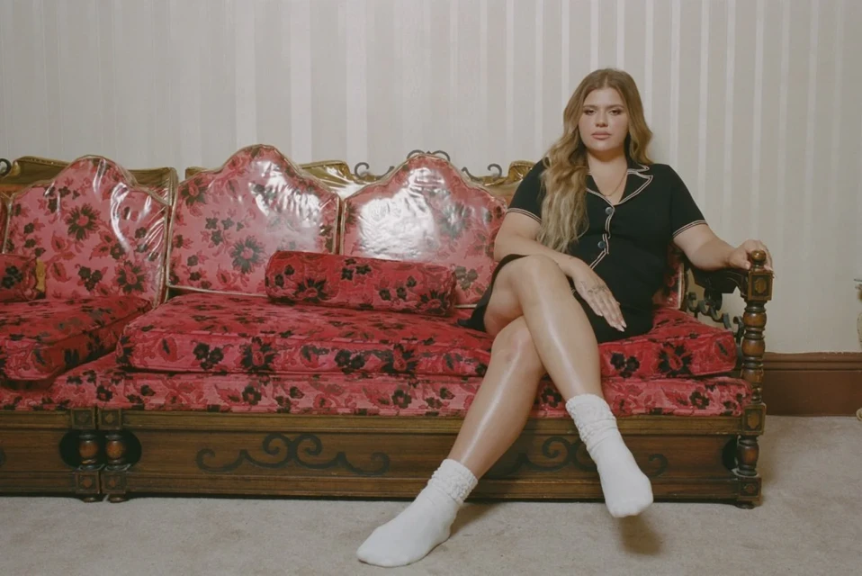 A woman with long blonde hair is sitting on a vintage red floral sofa, wearing a black dress and white socks, in an indoor setting.
