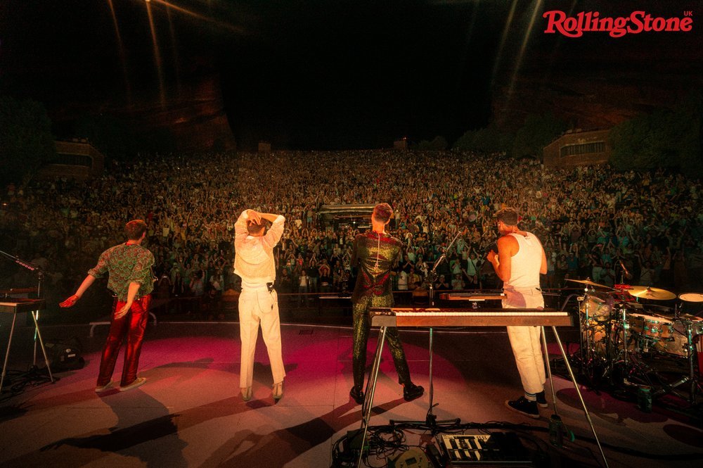Live concert performance featuring four musicians on stage performing for a large, cheering crowd in an outdoor amphitheater setting.