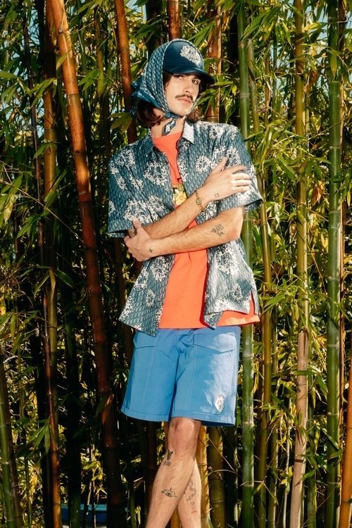 Adult male in an orange shirt and blue shorts stands in front of a bamboo forest wearing a patterned scarf and matching patterned shirt.