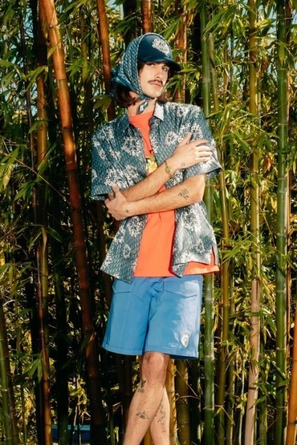 Adult male in an orange shirt and blue shorts stands in front of a bamboo forest wearing a patterned scarf and matching patterned shirt.