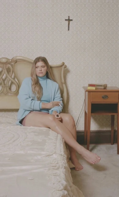 A woman with long blonde hair sits on a bed smoking a cigarette in a room with patterned wallpaper and a cross hanging on the wall.