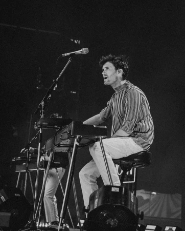 Monochrome shot of a male musician playing keyboard on stage at a concert, wearing a striped shirt and white pants, with a microphone visible.