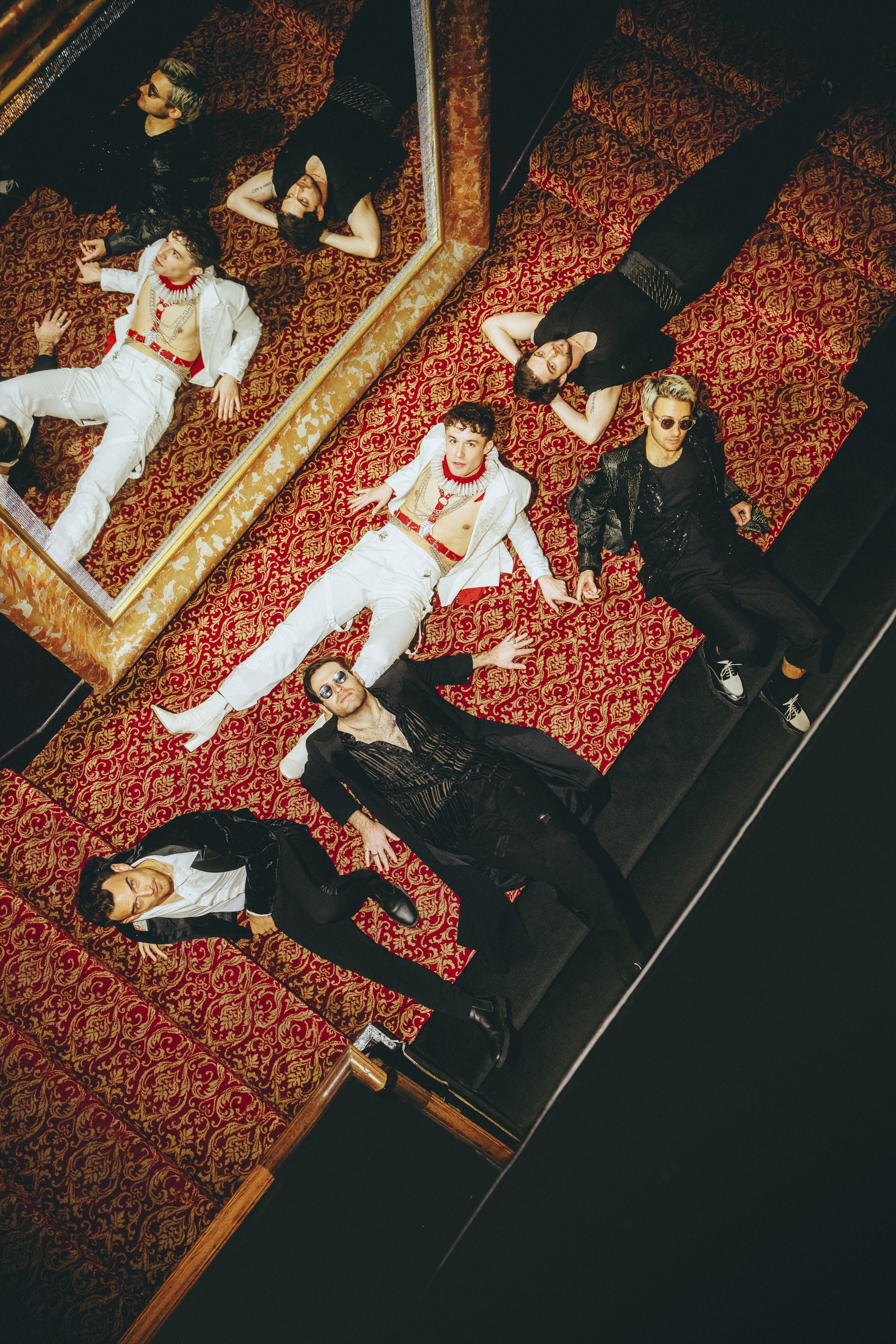 Group of seven people, male and female, wearing formal attire, laying on an patterned floor, shot from a high angle.