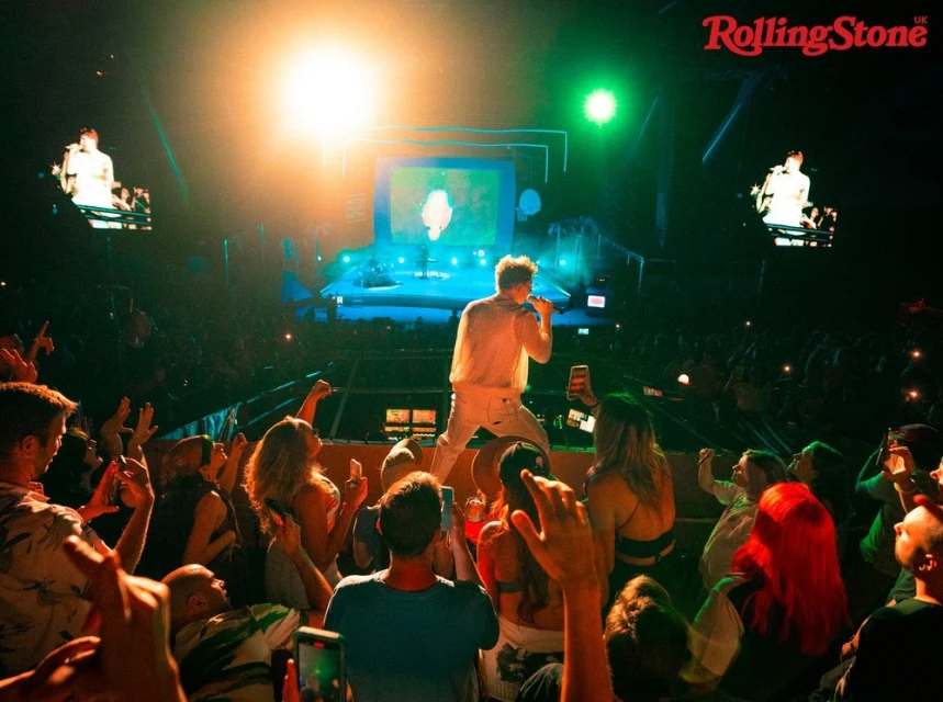 Singer performs on stage at a live concert in front of an enthusiastic crowd holding up their phones, illuminated by bright stage lights.