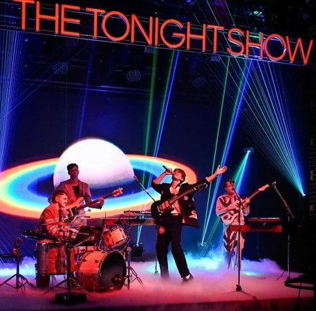 Band performs on stage for The Tonight Show, featuring musicians with instruments, stage lights, and fog effects in a concert setting.