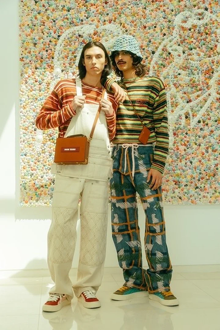 Two fashion models in striped tops and patterned pants pose indoors in front of a textured wall, showcasing unique style and design.