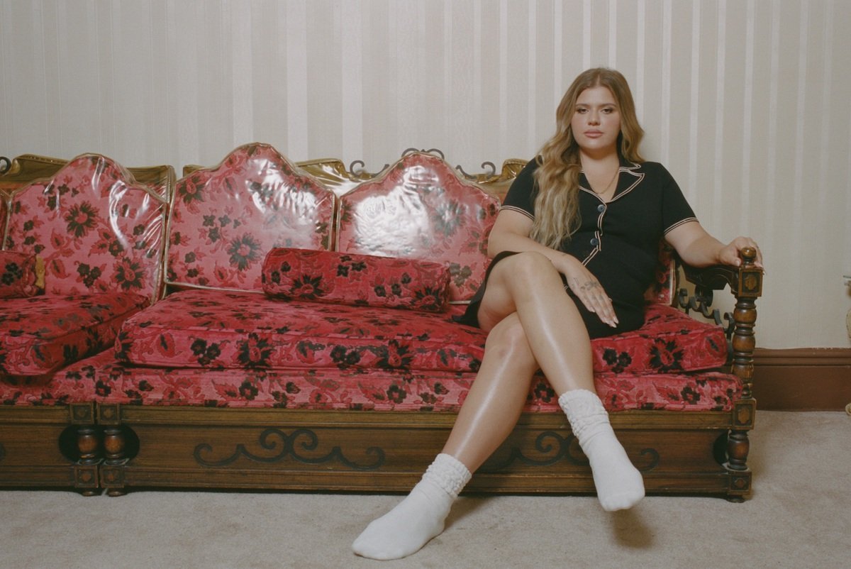 Woman in black dress and white socks sits on a red floral couch in an indoor setting, captured in eye-level shot.