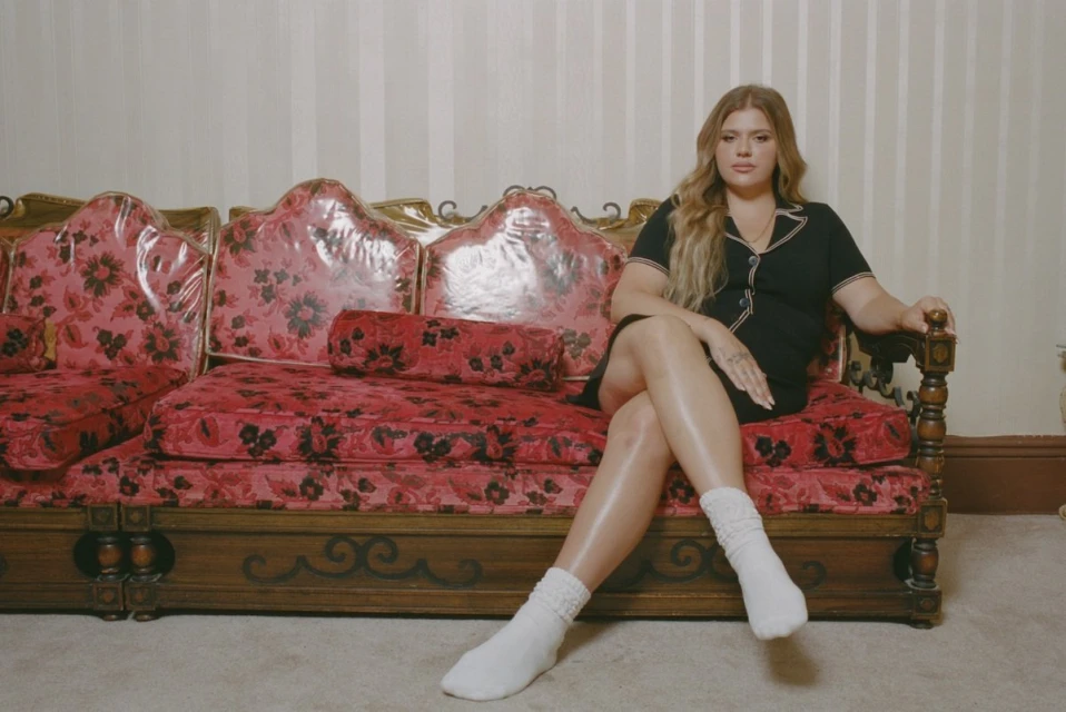 Woman in black dress and white socks sits on a red floral couch in an indoor setting, captured in eye-level shot. 