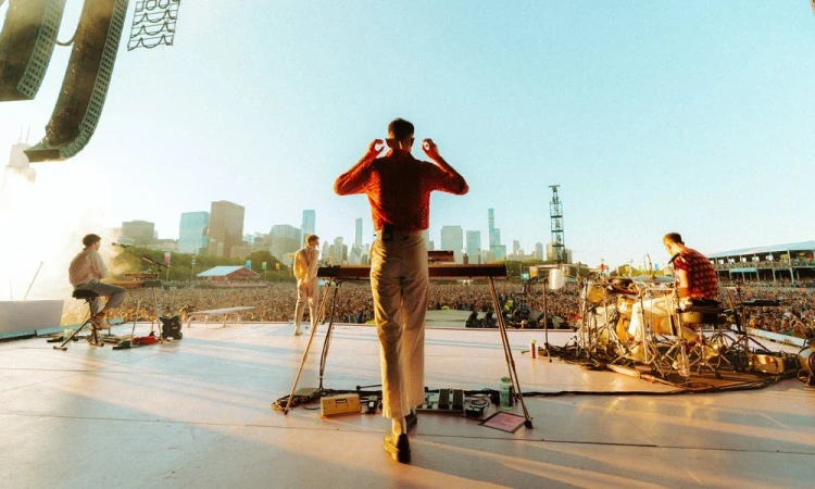 Band performs on outdoor stage at music festival with city skyline backdrop, drawing a large crowd in daytime concert setting.