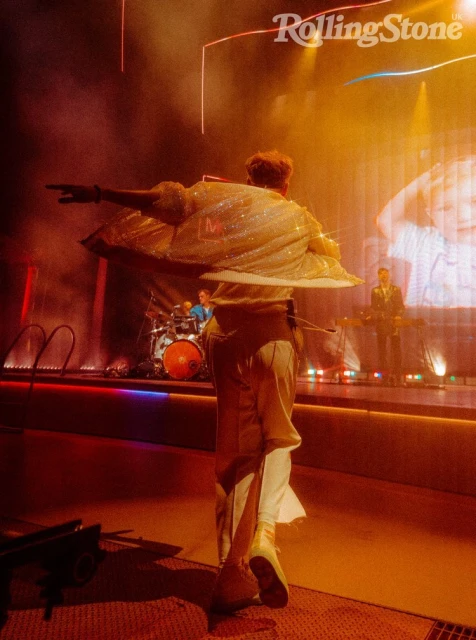 Back view of a male musician with outstretched arm, wearing a sparkly jacket and white pants, performing onstage with his band under orange lights.
