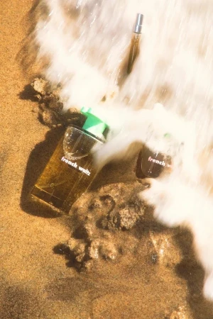 Three 'French Waltz' perfume bottles from Tyler, the Creator on sandy beach with soft, white waves washing up around the base of the bottles.