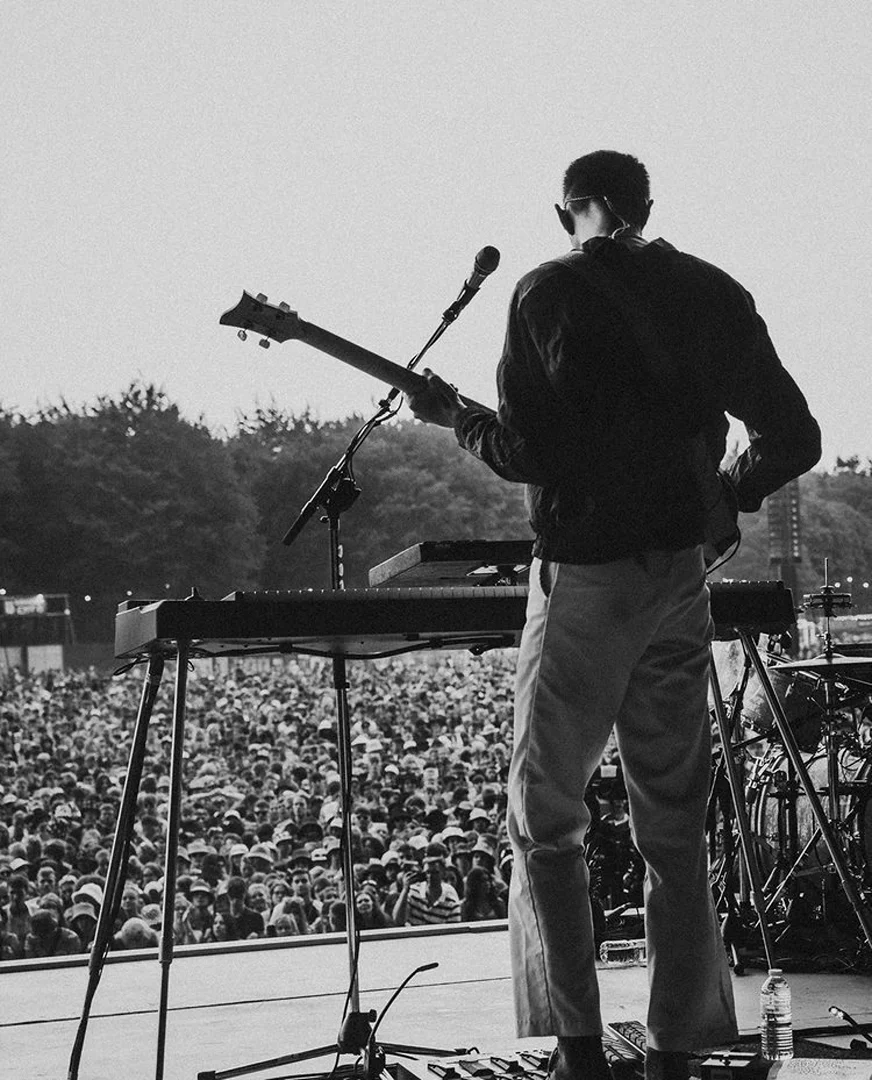 Monochrome shot of a musician playing guitar and keyboard on stage during an outdoor concert, facing away from the camera towards a crowd.