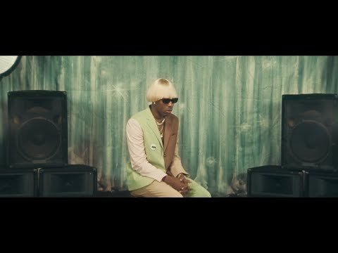 A man with blonde hair and sunglasses is sitting between two black speakers in front of a green patterned wall, looking thoughtful.