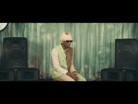 A man with blonde hair and sunglasses is sitting between two black speakers in front of a green patterned wall, looking thoughtful.