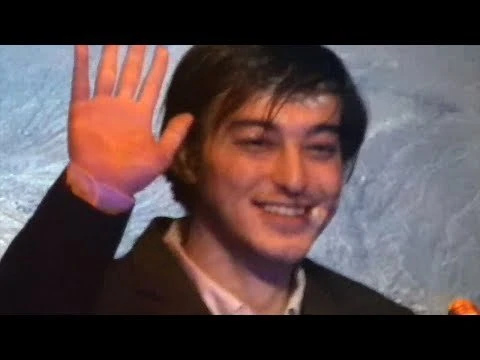 A smiling adult male raises a hand in a light-filled indoor scene, wearing a dark jacket against a textured light blue background.