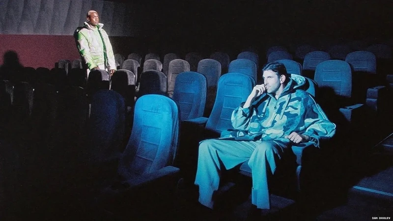 Two men in an empty blue movie theatre, one standing in a lime green coat, one sitting in a camouflage jacket.