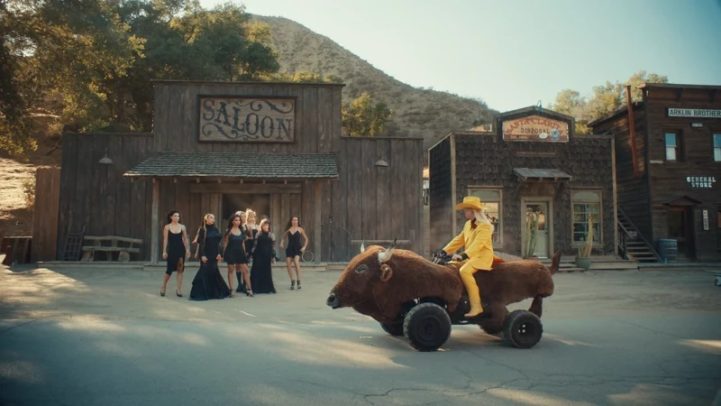 A person in a yellow suit rides a faux-buffalo vehicle on a Western town street with women in black dresses standing nearby a saloon.