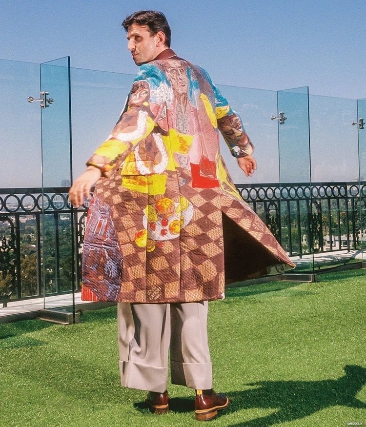 Adult male poses outdoors wearing an artistically patterned overcoat, tan pants, and brown shoes, against a green grass and urban backdrop.