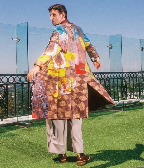 Adult male poses outdoors wearing an artistically patterned overcoat, tan pants, and brown shoes, against a green grass and urban backdrop.