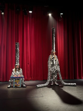 Two performers in shiny silver costumes and tall headwear pose on a stage with red curtains, lit by a spotlight, creating a surreal scene.