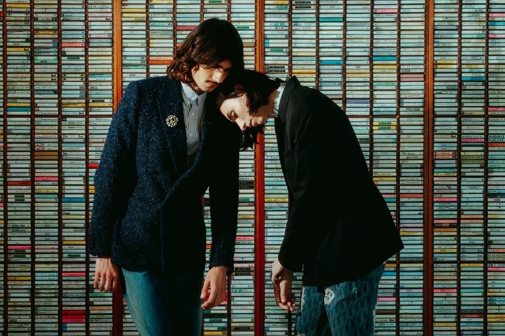 Two people pose indoors in dark blazers with a wall of cassette tapes as a backdrop, creating a retro, music-themed fashion shot.
