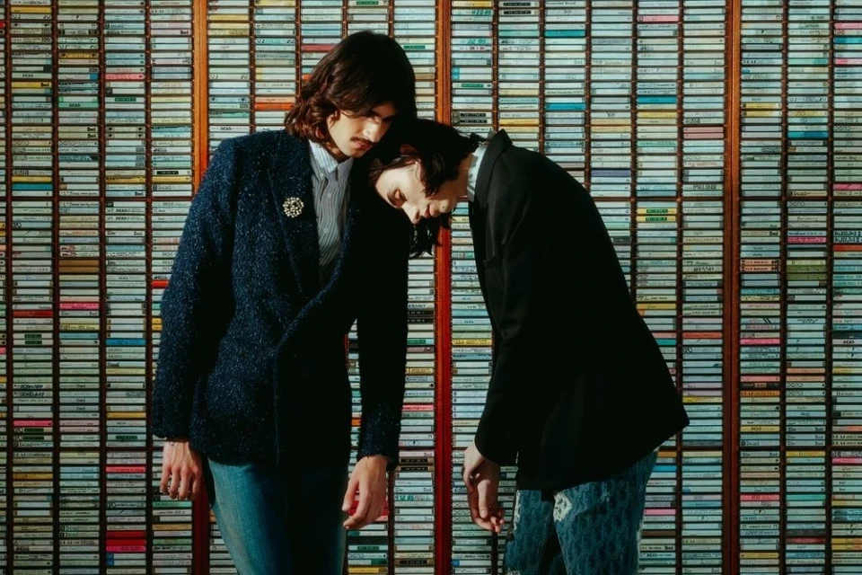 Two people pose indoors in dark blazers with a wall of cassette tapes as a backdrop, creating a retro, music-themed fashion shot.