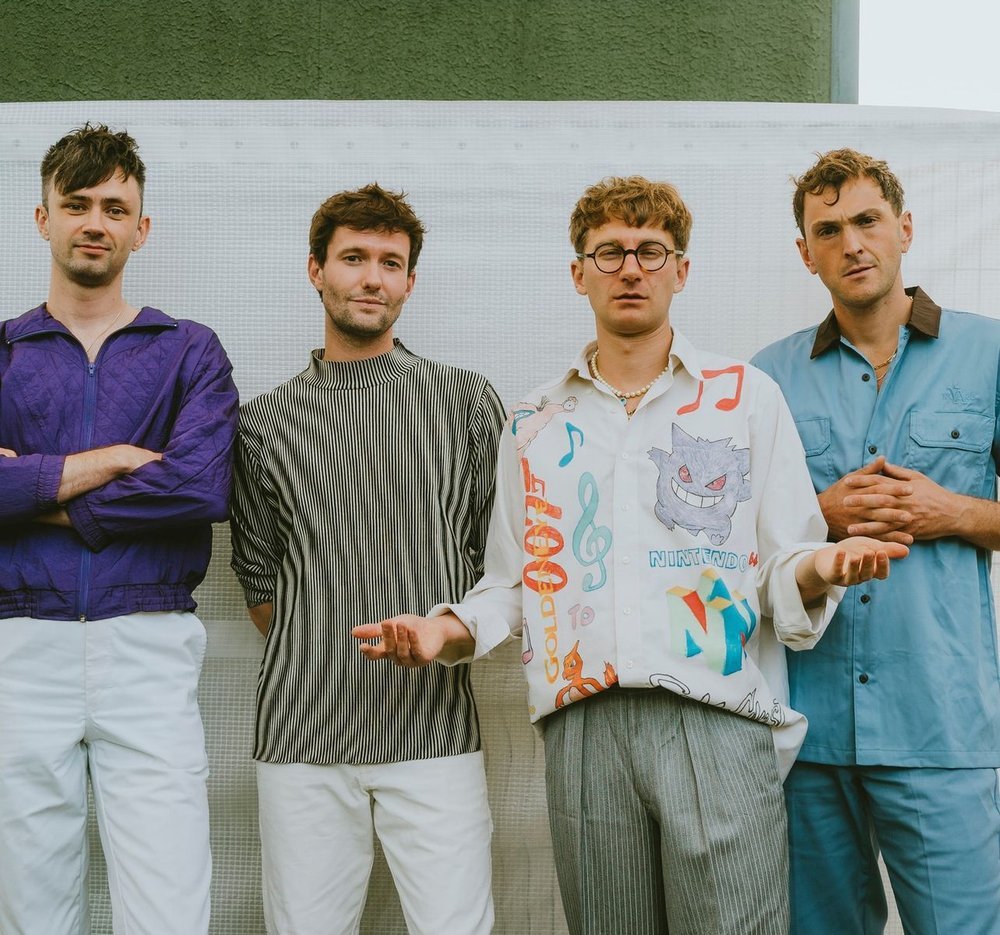 Group of four young adult men posing indoors against a white background, wearing casual colorful clothing and neutral pants.