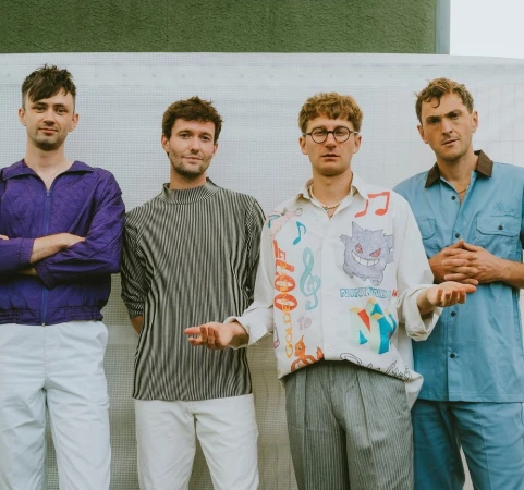 Group of four young adult men posing indoors against a white background, wearing casual colorful clothing and neutral pants.