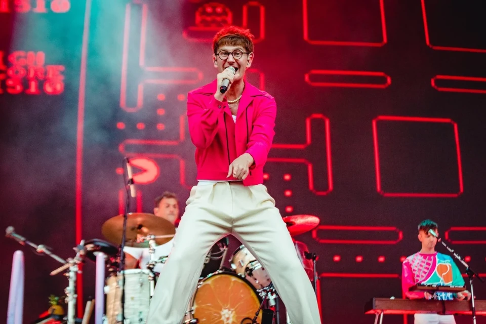 A performer sings on stage in a pink jacket with a band in the background and stage lighting for a music performance.