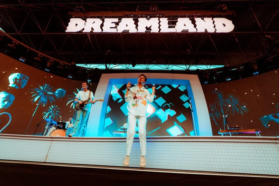 Band performs live on stage at Dreamland venue. Singer in white suit stands in front of screen; guitarist, drummer, and keyboardist visible.
