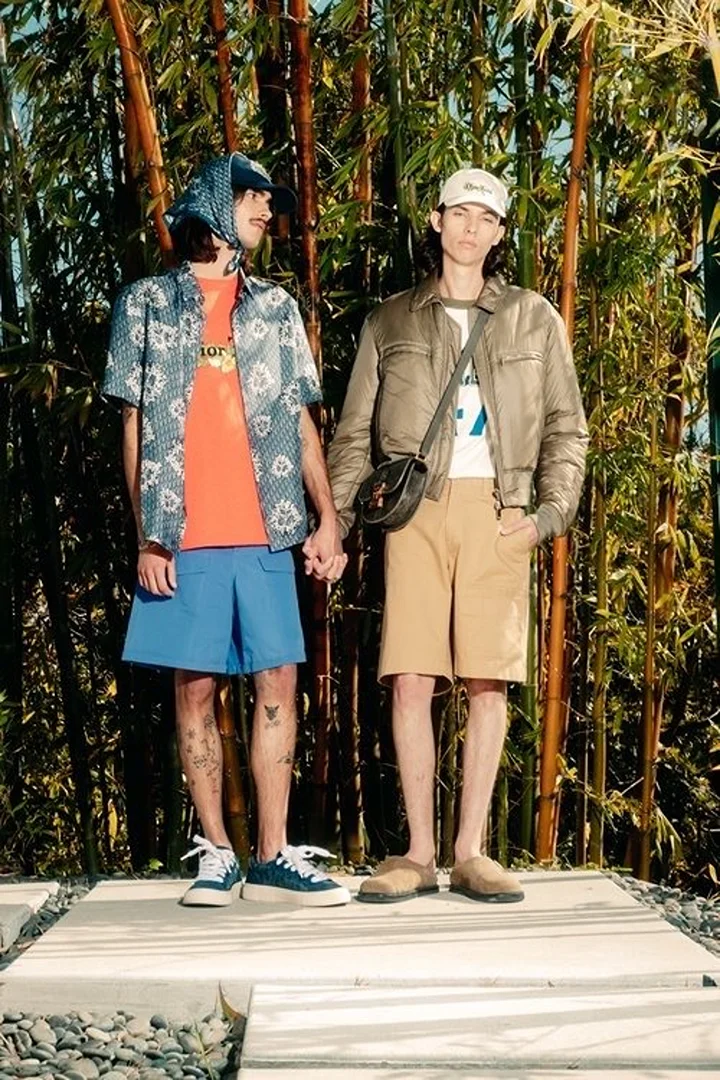 Two adult males holding hands in front of bamboo. One wears a headscarf, orange shirt, and blue shorts. The other wears khaki shorts and a jacket.