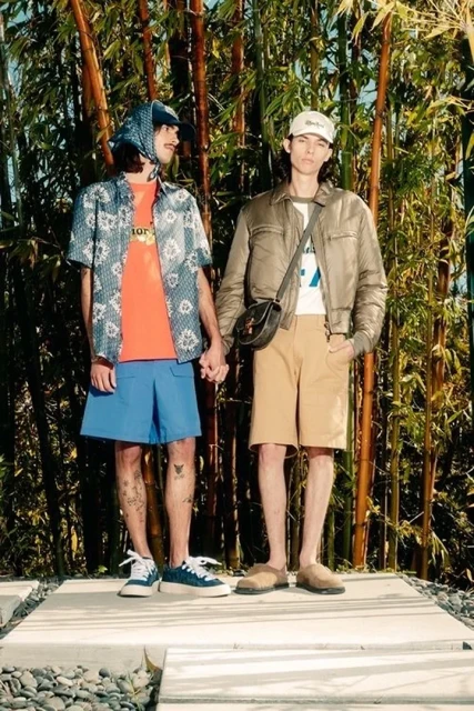 Two adult males holding hands in front of bamboo. One wears a headscarf, orange shirt, and blue shorts. The other wears khaki shorts and a jacket.
