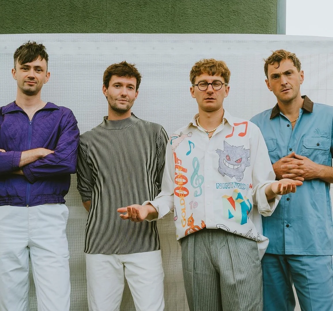 Four young adult men stand in a row, wearing fashionable shirts and pants. One man wears glasses and the others stand beside him.